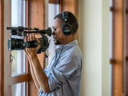 Student from 传播与媒体学院 wear headphones and holding a video camera filming through a window.